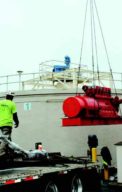 A red industrial machine is being hoisted by a crane, with a worker in a safety helmet supervising the operation.