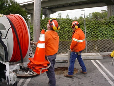 Two workers in orange safety gear with helmets and ear protection, near equipment and an open manhole on a road.