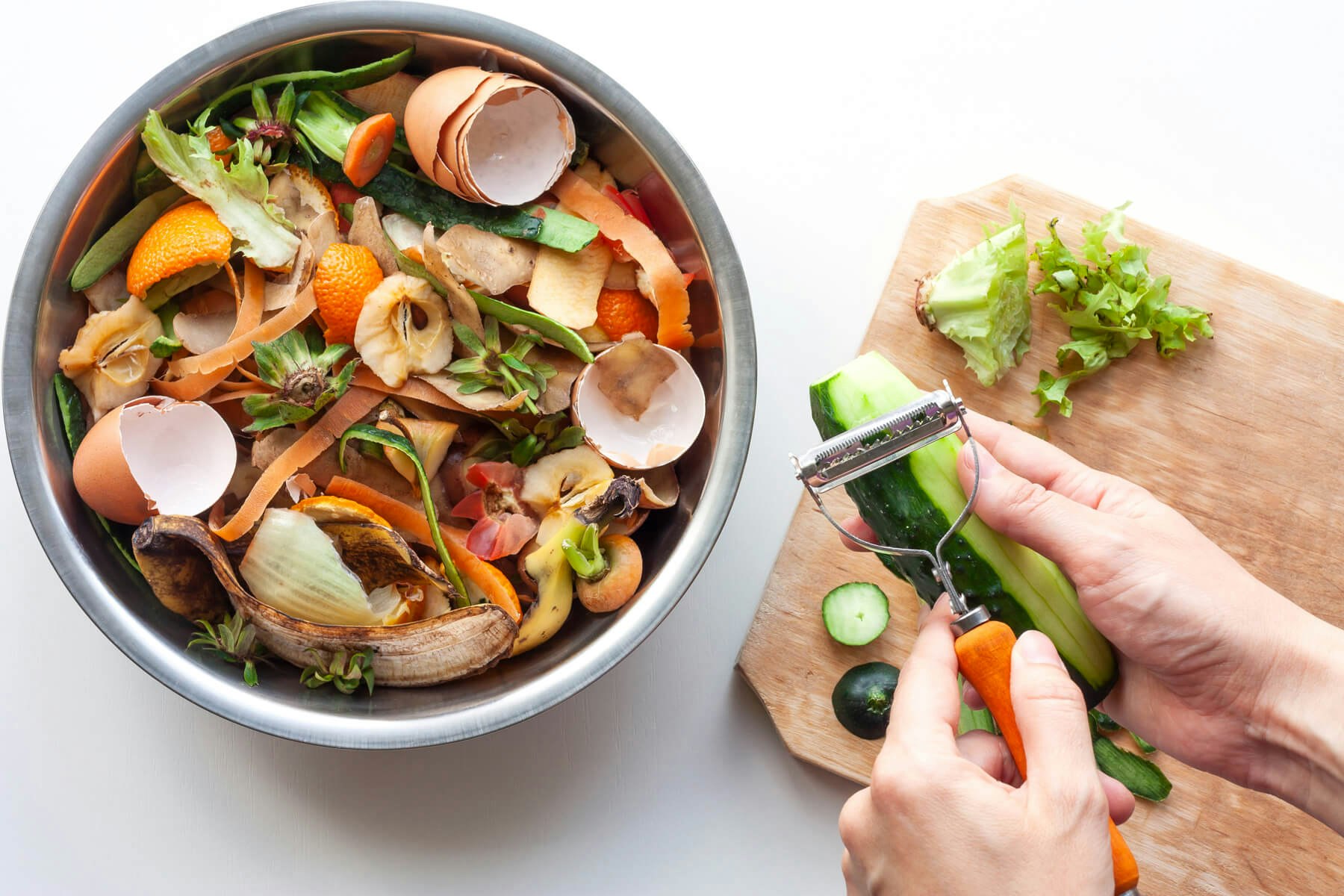 A bowl of food scraps including eggshells, fruit peels, and vegetables, next to hands peeling a cucumber.
