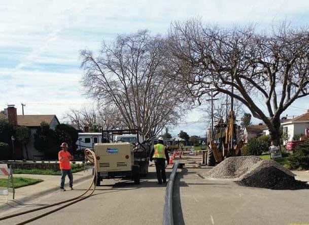 Construction workers in bright vests working on a residential street with machinery and gravel piles, alongside bare trees.
