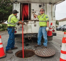 Two workers in safety vests manage equipment near an open manhole, with traffic cones around them.