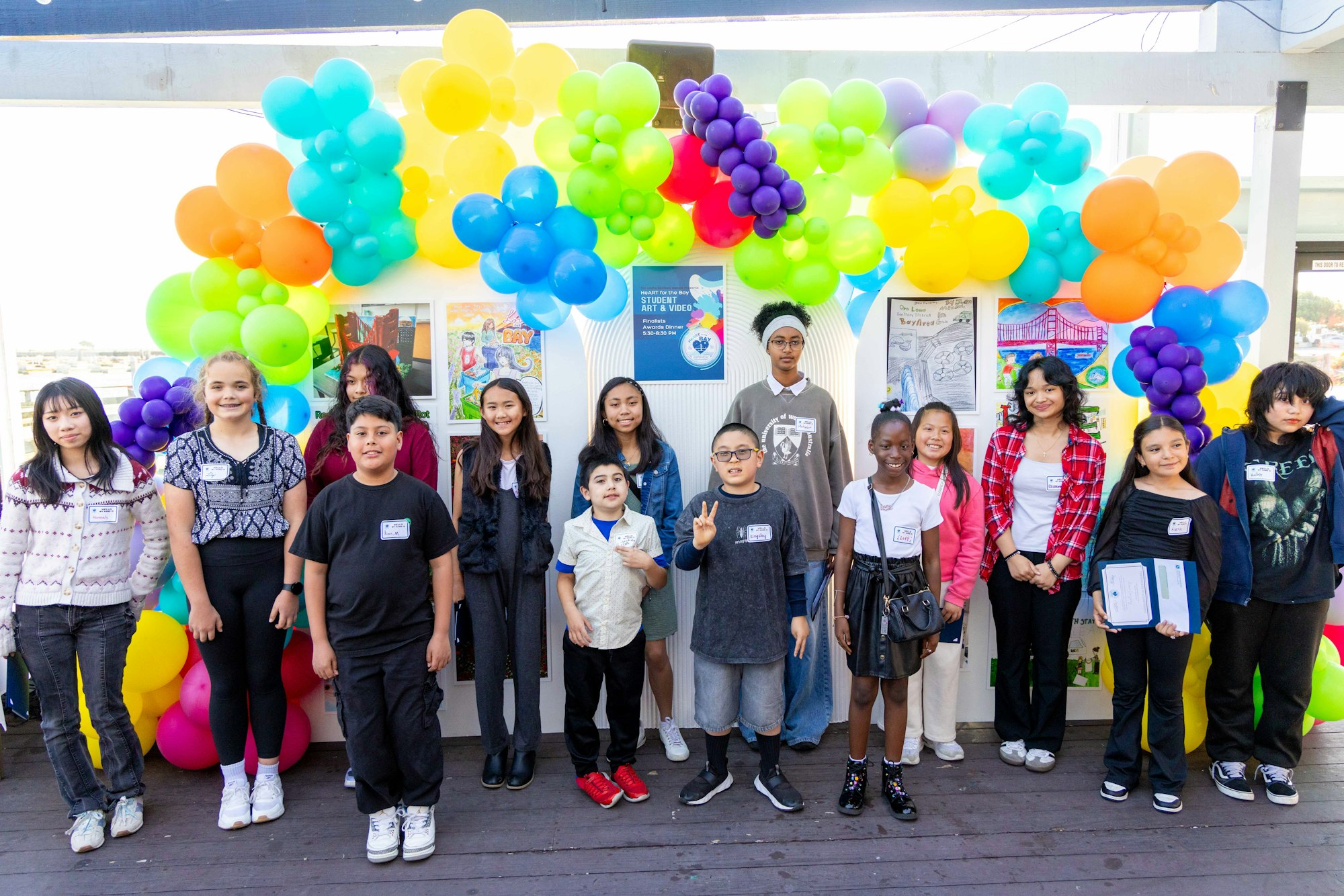 A group of kids poses together in front of colorful balloon decorations at an art event showcasing their work.
