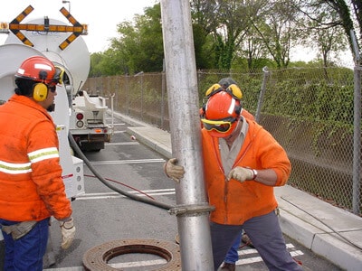 Workers in safety gear handling a large pipe near a manhole and utility truck on a street.