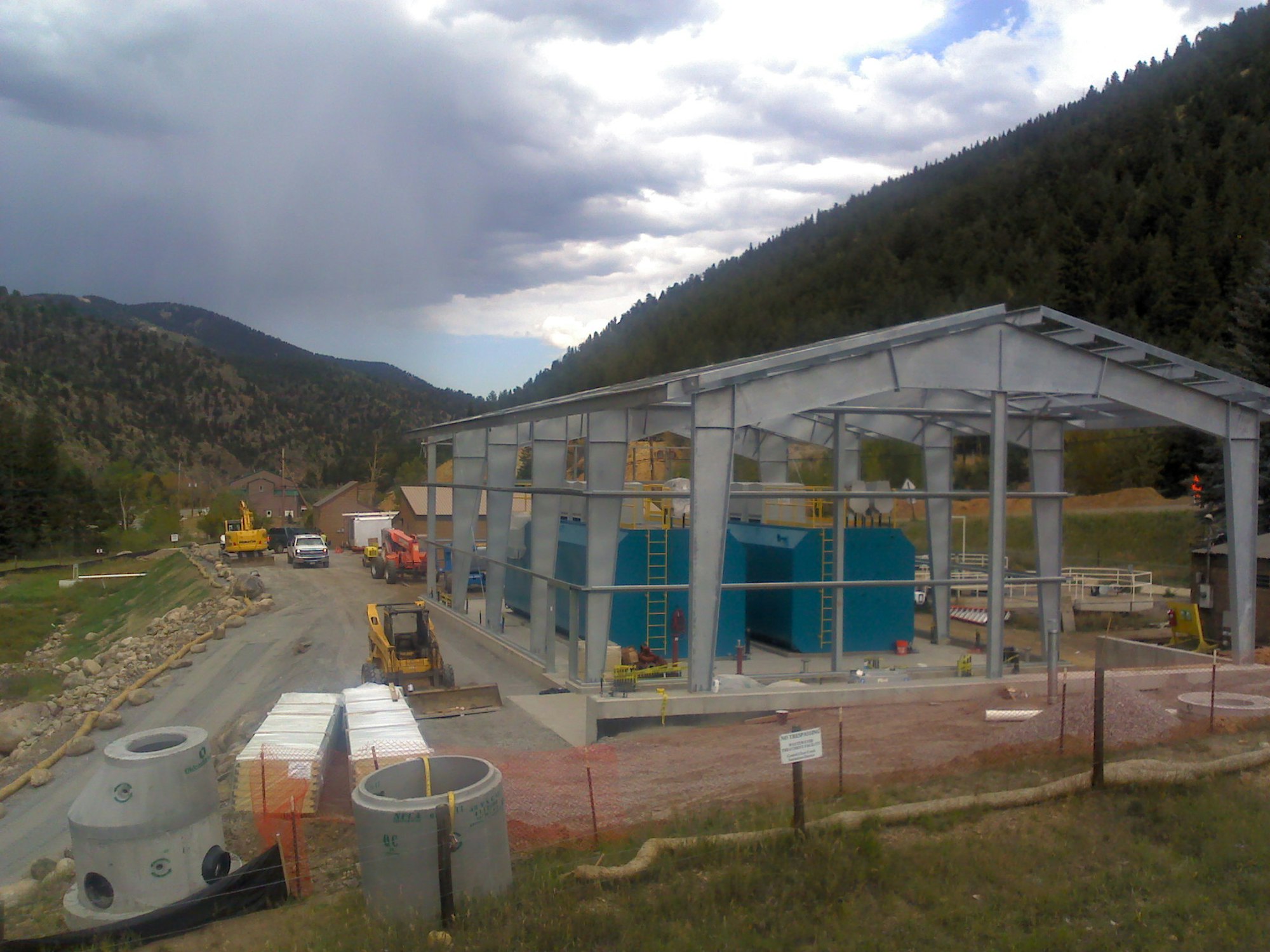 A construction site with a steel framework for a building and industrial equipment, surrounded by mountains.