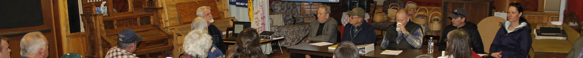 People in a wood-paneled room, possibly at a meeting or community event, sitting and facing forward.