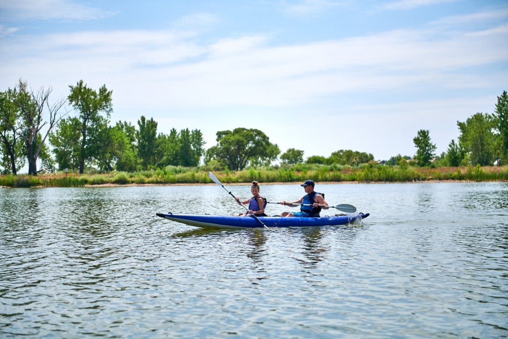 Two people kayaking on a calm lake with trees and sky in the background.