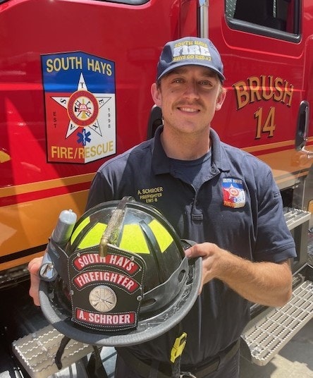 A firefighter stands in front of a fire truck, holding a helmet labeled "South Hays Firefighter A. Schroer."