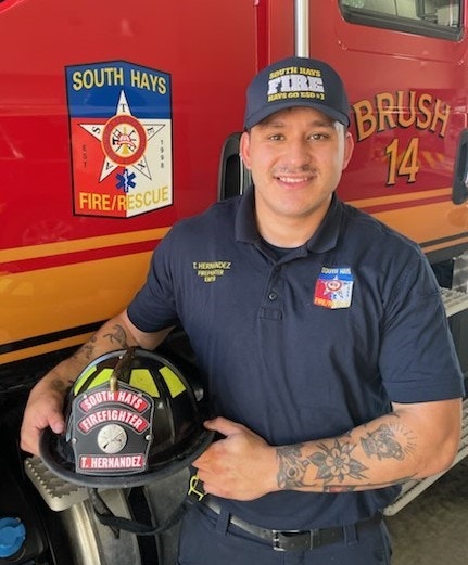 A firefighter in uniform stands next to a fire truck, holding a helmet labeled "South Hays Firefighters."