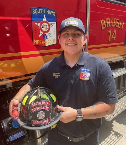 A firefighter smiling, holding a helmet, in front of a red fire truck labeled "BRUSH 14" with a "South Hays Fire/Rescue" emblem.