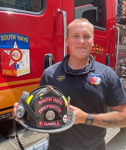A firefighter in uniform holds a helmet in front of a fire truck with "South Hays Fire/Rescue" logos.