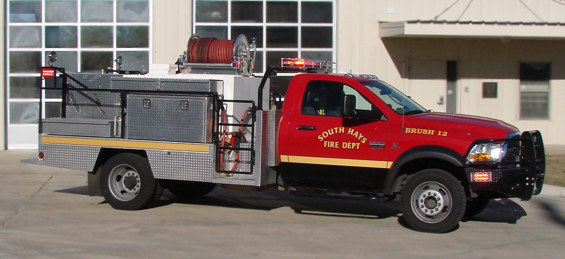 A red and silver fire department brush truck with lights on, parked outside a building.