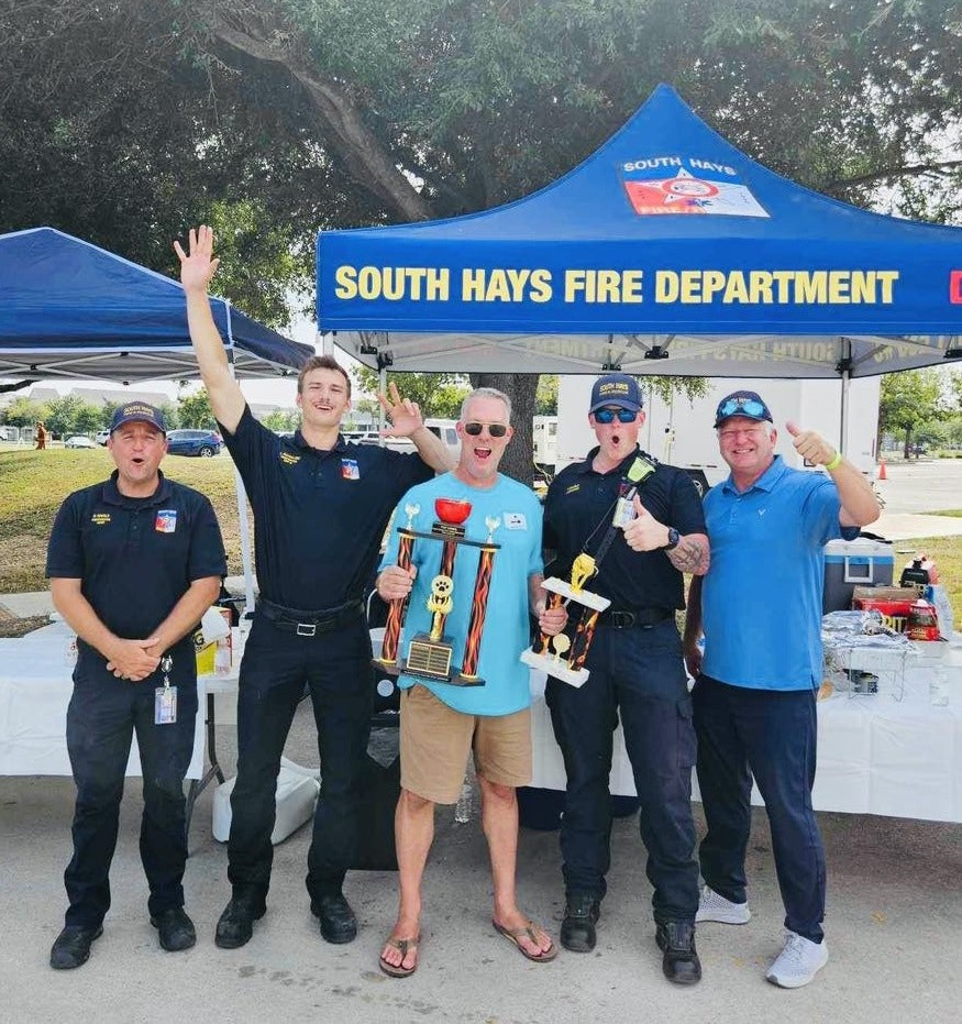 Group of happy people with trophies in front of a South Hays Fire Department tent.