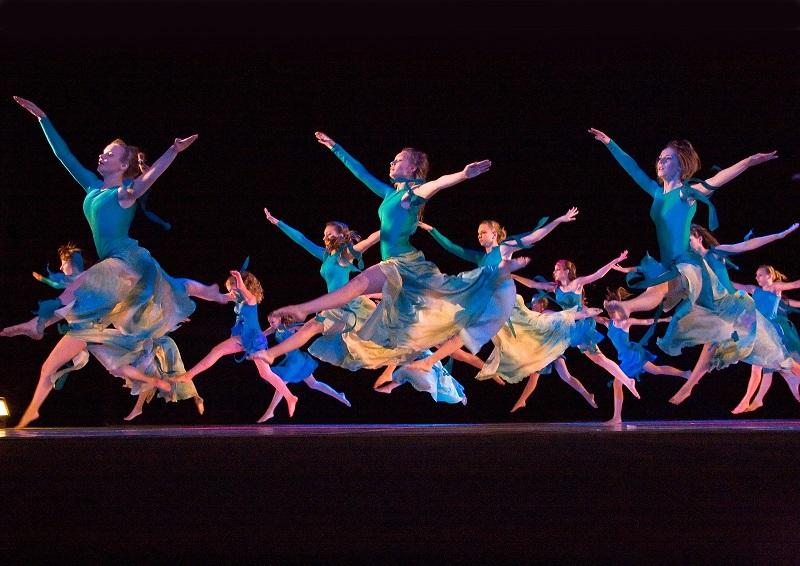 A group of dancers in flowing teal costumes performing an energetic jump on stage, showcasing grace and synchronization.