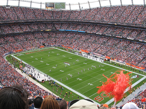 A large crowd fills a stadium, watching a football game on the field below, with fans waving pom-poms and cheering.