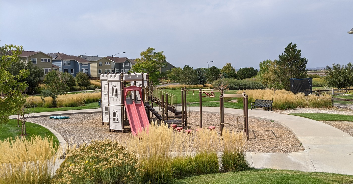 A playground featuring a castle structure with a slide, surrounded by grass, benches, and residential buildings in the background.