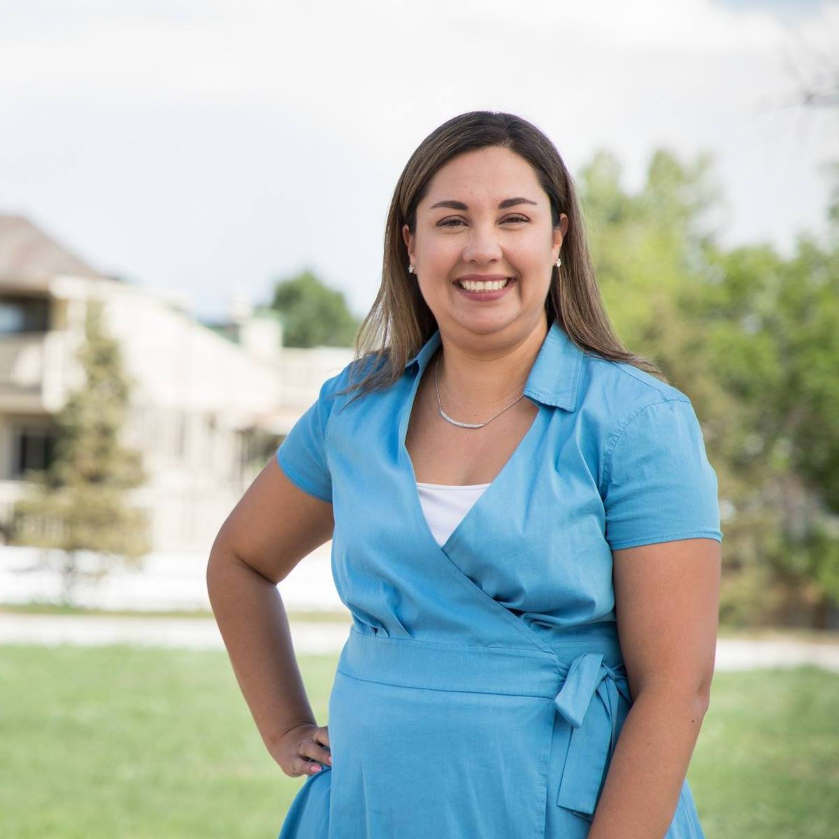 A smiling woman in a light blue dress stands outdoors, with green grass and trees in the background.