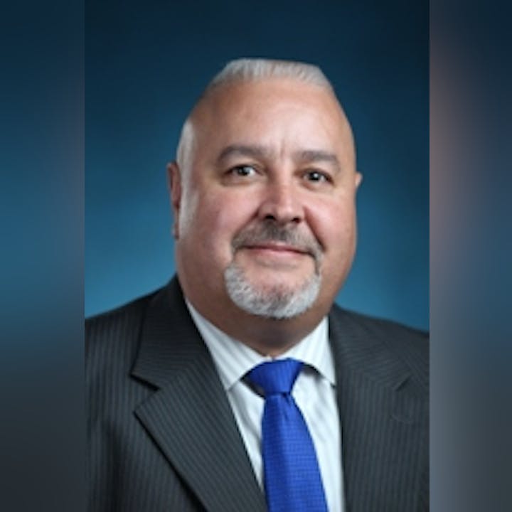 The image features a man in a suit and tie, smiling against a blue background. He has short, gray hair and a goatee.