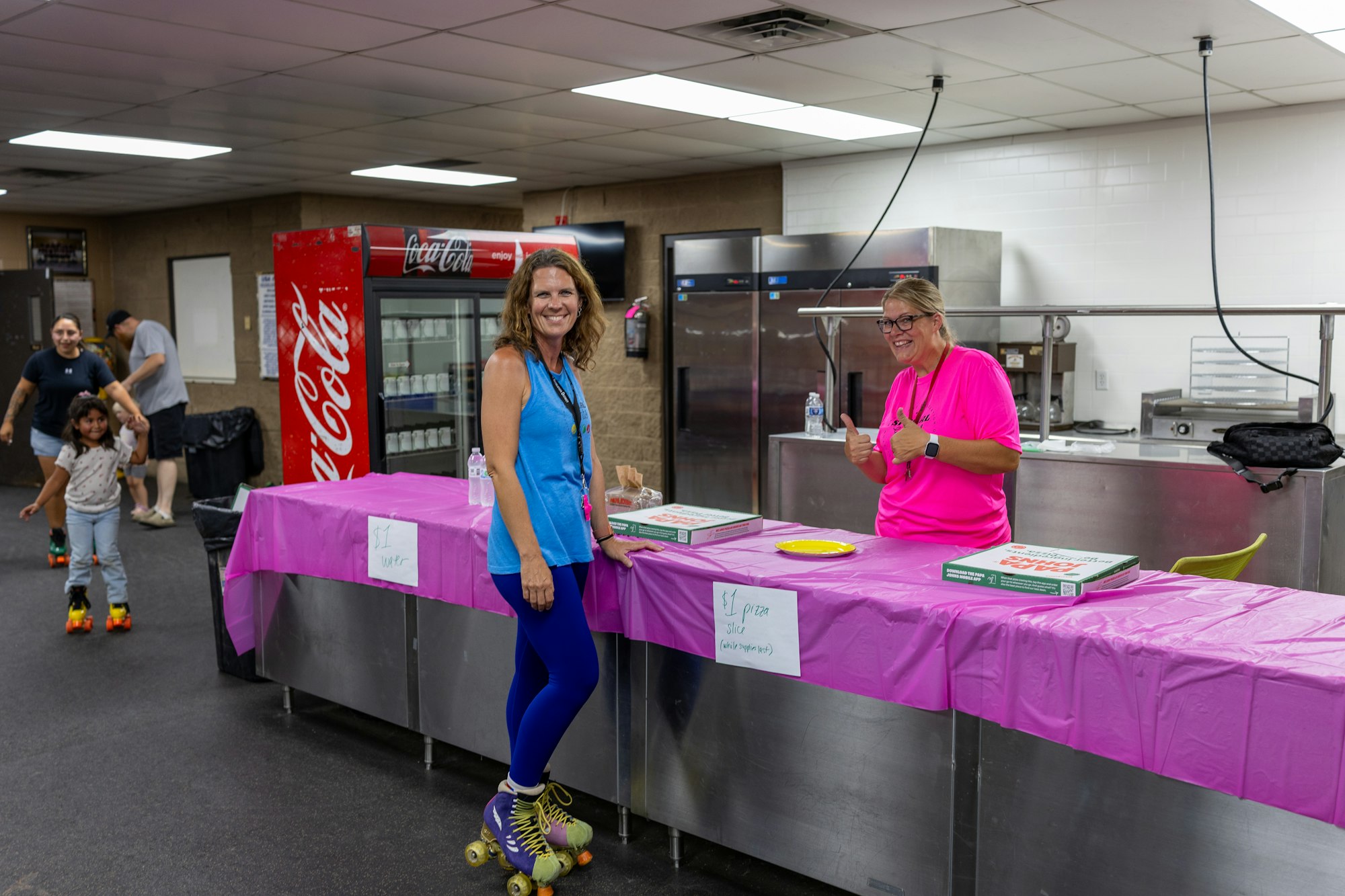 Two people on roller skates near a counter selling snacks. Pizza for $1/slice, water for $1. People skating in the background.