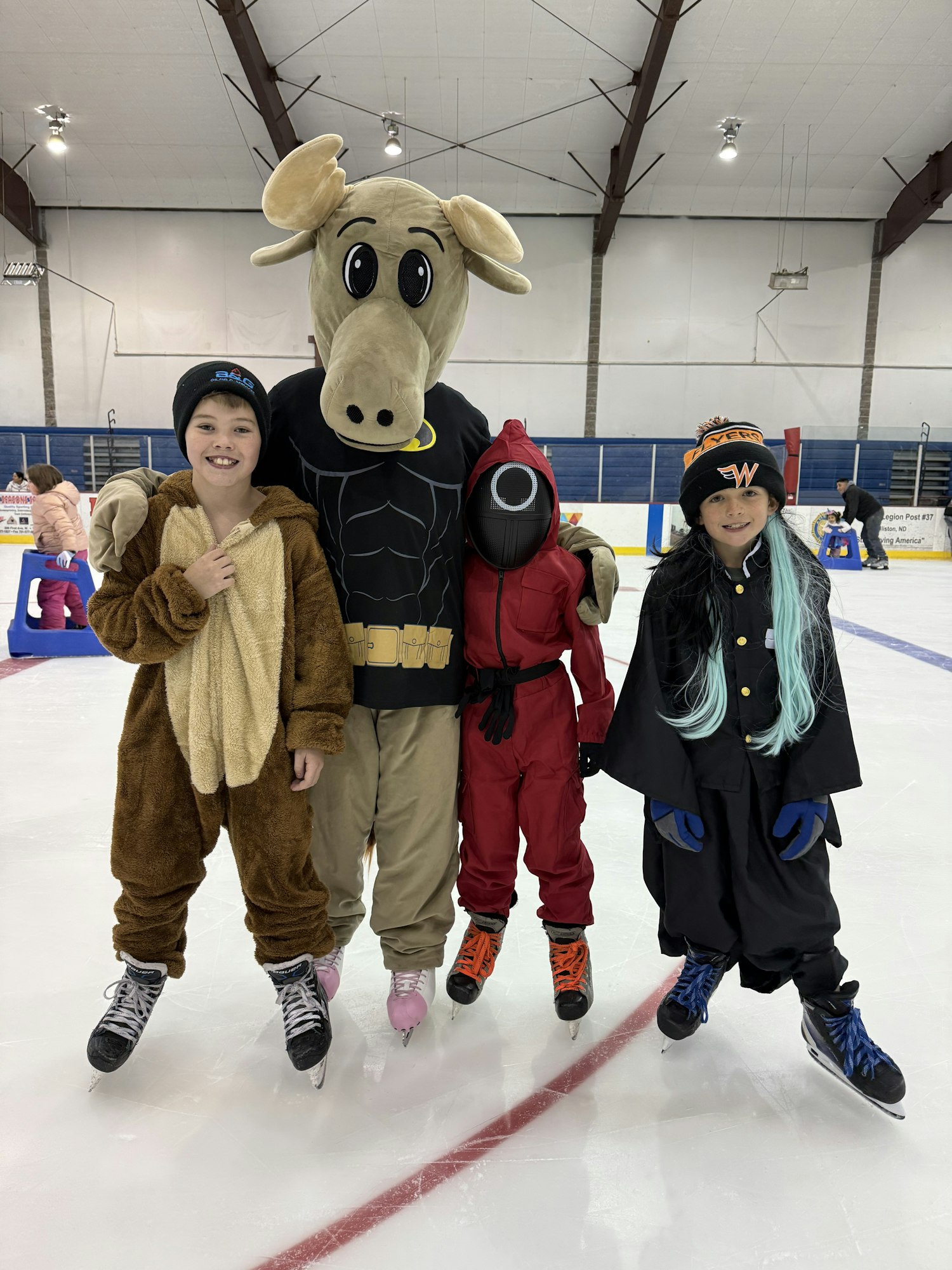 A group of kids on an ice rink, dressed in costumes, including a moose mascot, colorful outfits, and ice skates.