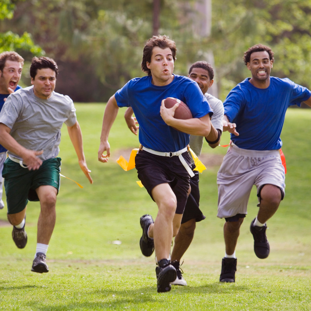 A group of men are playing flag football in a park, with one runner holding the football while others chase him.