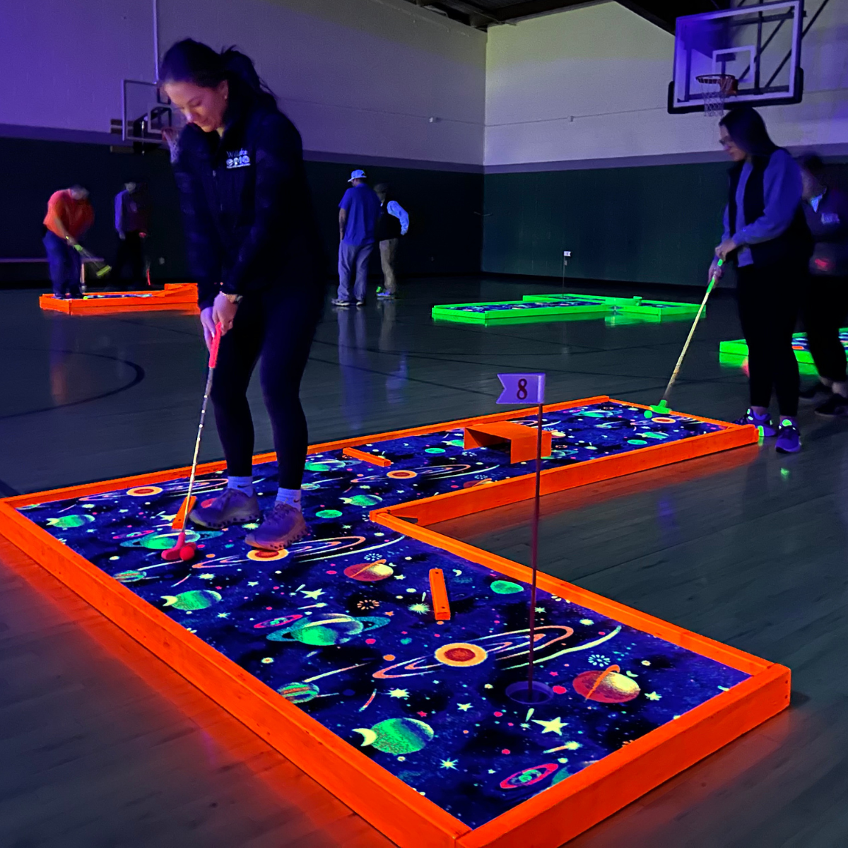 People playing glow-in-the-dark mini-golf indoors with space-themed designs.