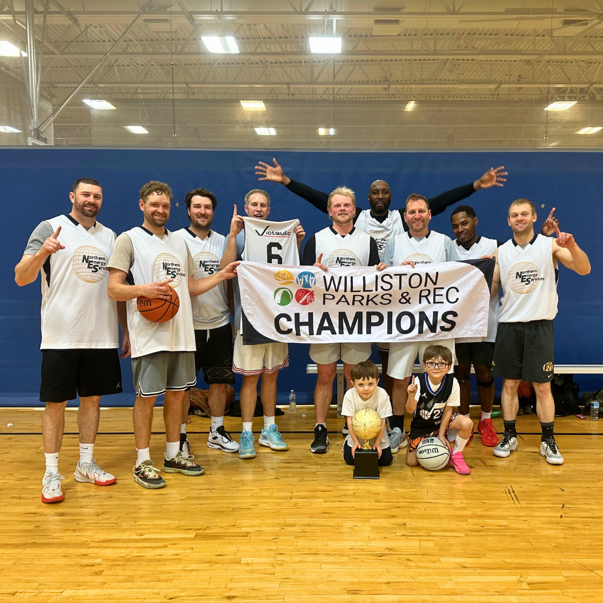 A basketball team celebrates their victory as champions, holding a banner and trophy, with a couple of kids in the front.