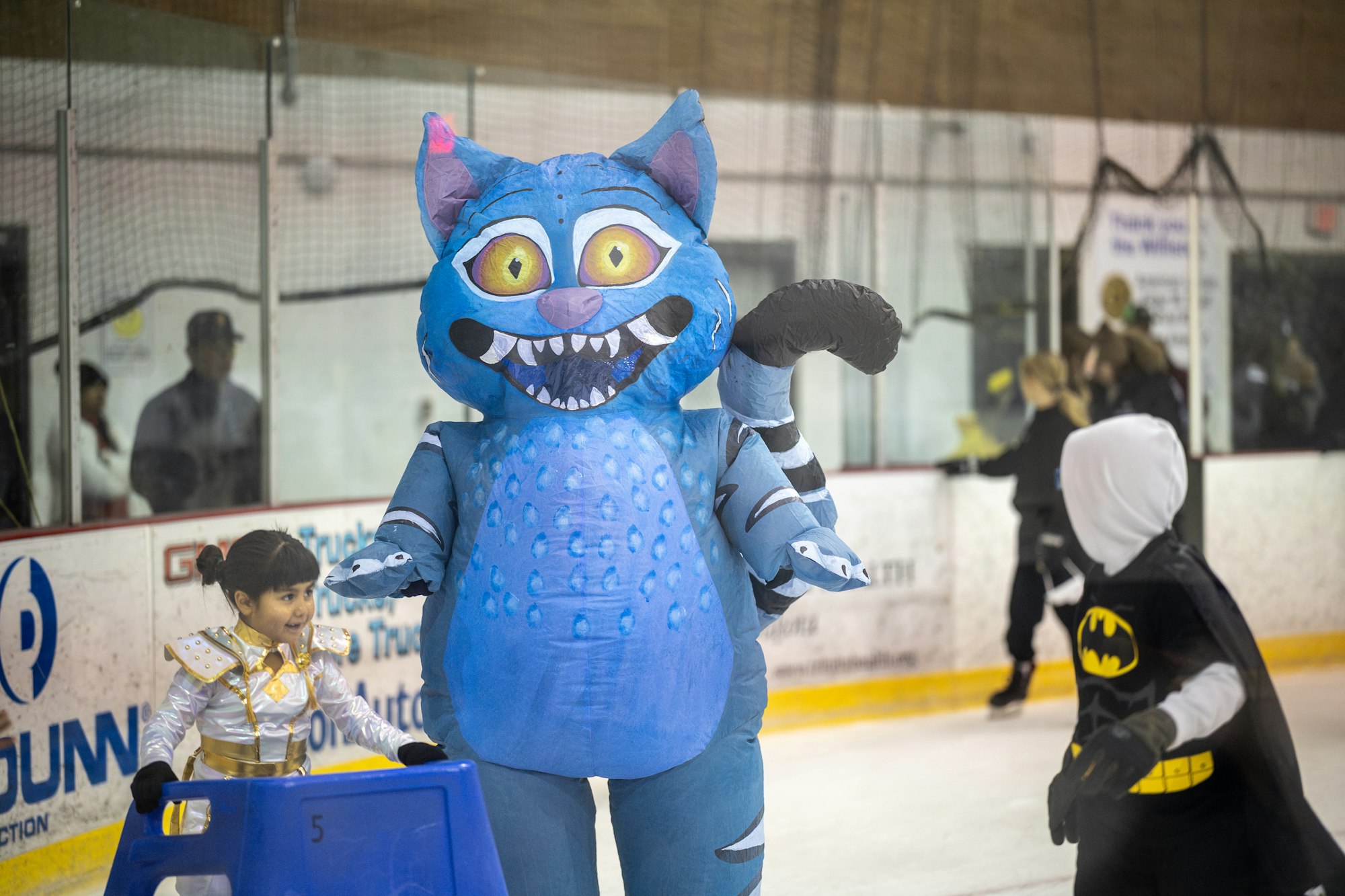 A blue costumed character skates with a child dressed as a princess on ice, while another child in a Batman costume watches.