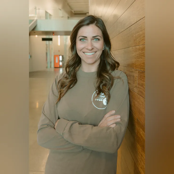 A woman with long hair smiles confidently, wearing a brown sweatshirt, standing against a wooden wall in a well-lit space.