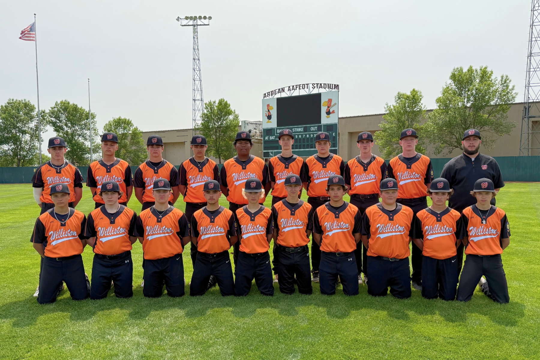 A baseball team in orange "Williston" jerseys poses on a field in front of a stadium scoreboard.