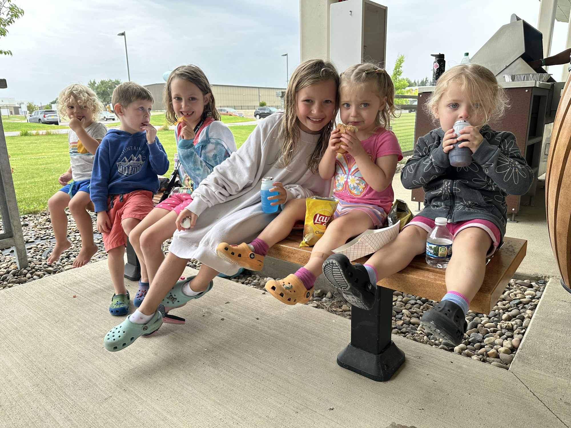 Group of children sitting outdoors, some smiling, with snacks and drinks.