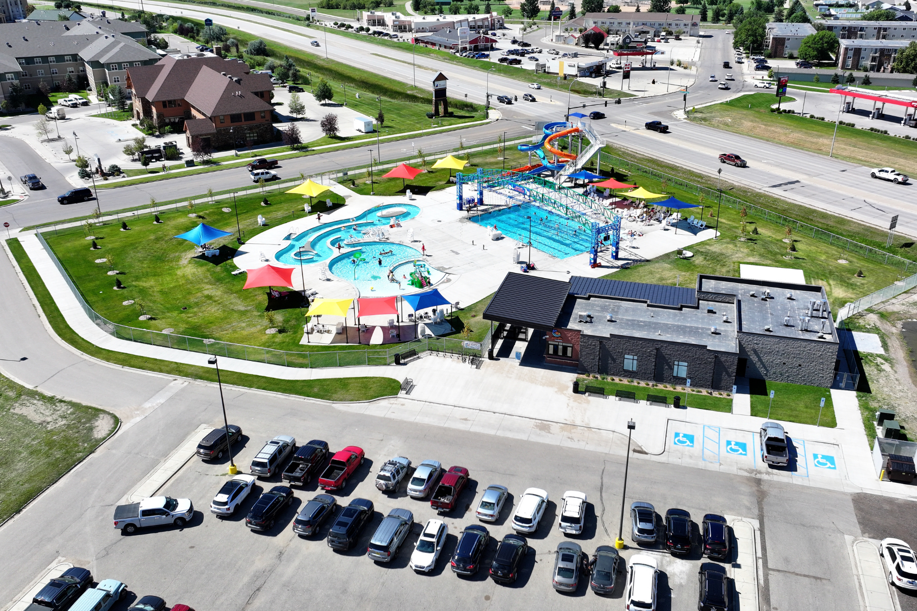 The image shows a bustling outdoor swimming pool area with water slides, colorful tents, and a parking lot nearby.