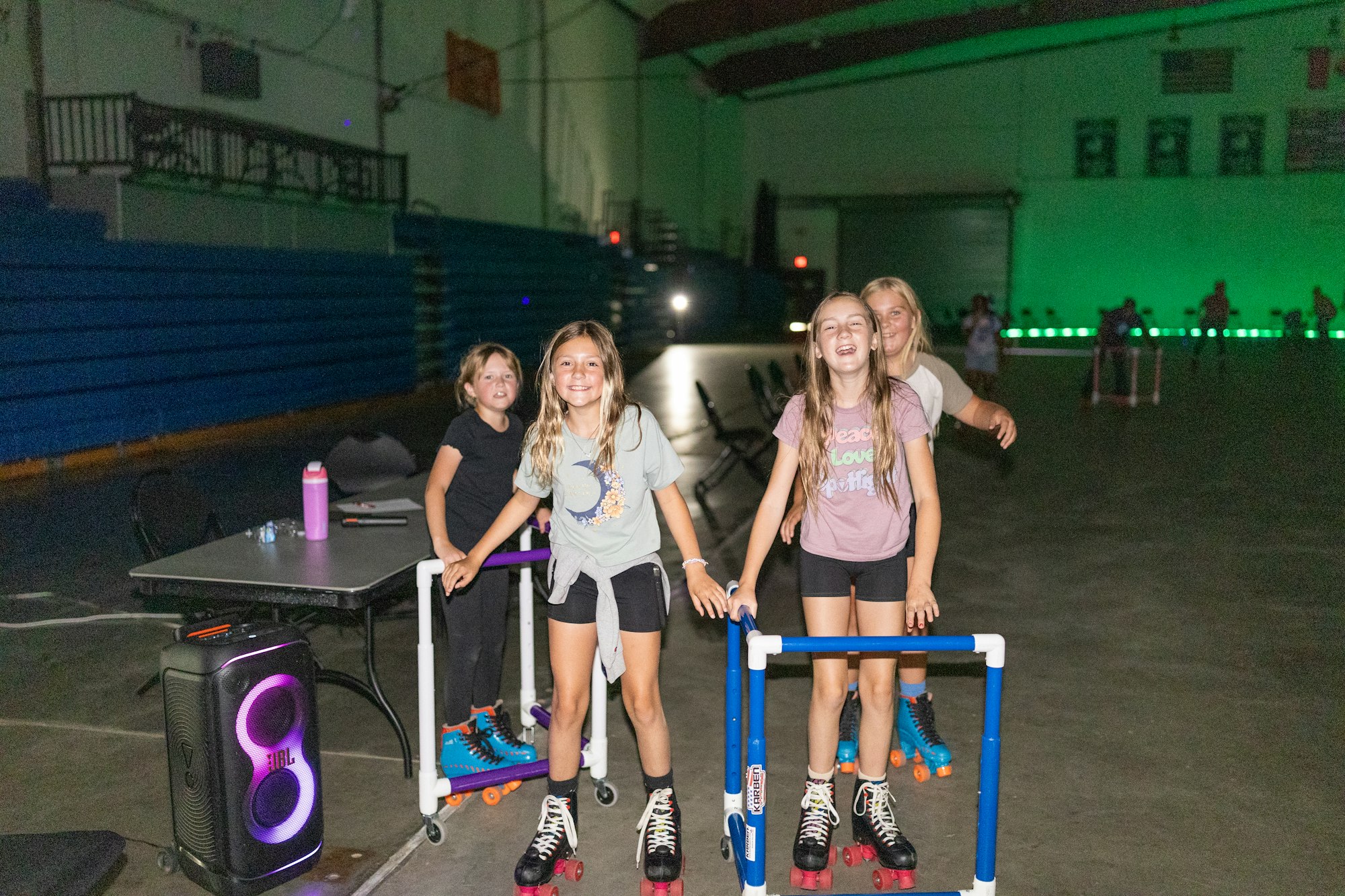 Four kids roller skating indoors, smiling and using support bars, with a speaker and table nearby.