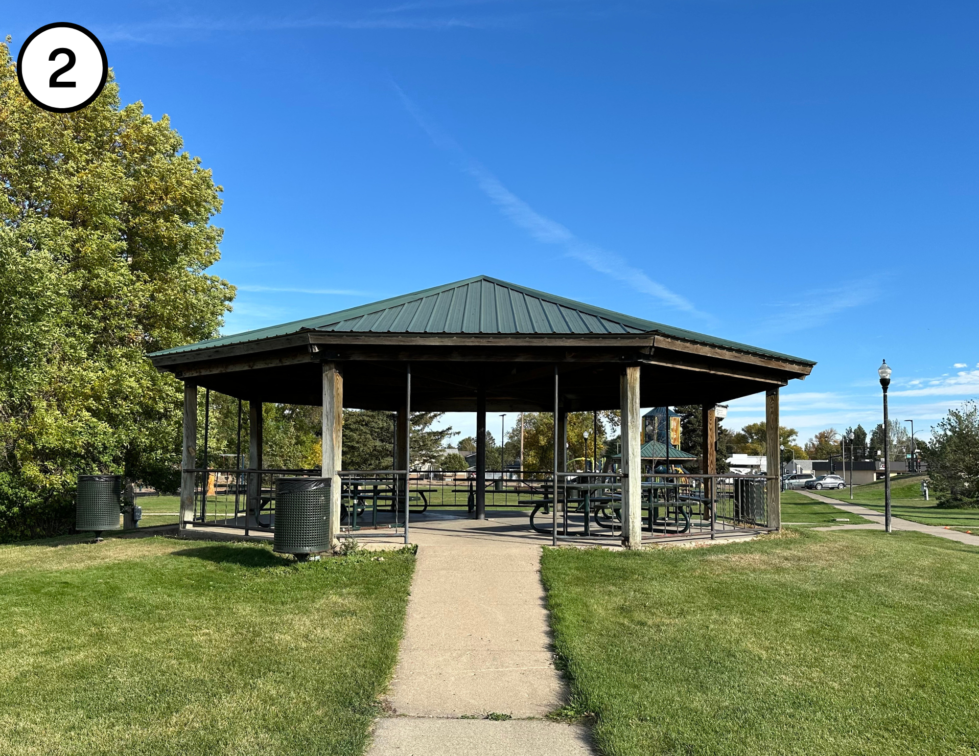 A gazebo with a metal roof in a park, surrounded by grass and trees, with a clear blue sky.