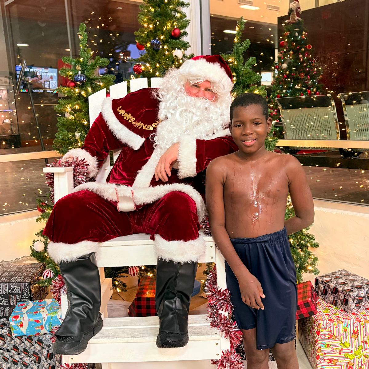 A boy in swim trunks stands next to Santa Claus, who is seated amidst festive decorations and Christmas presents.