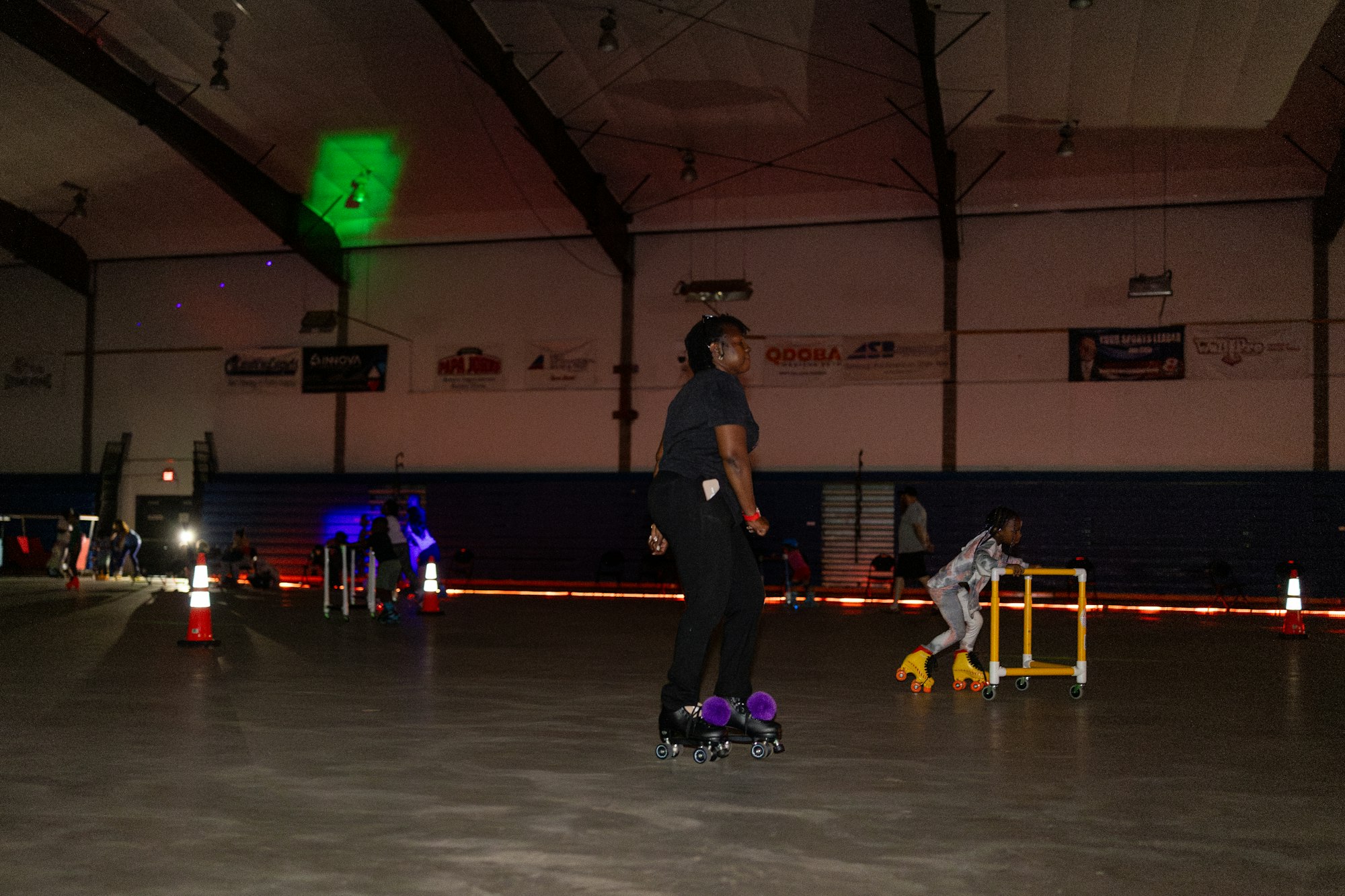 People roller skating indoors, dim lighting, colorful skates, cones on the floor.
