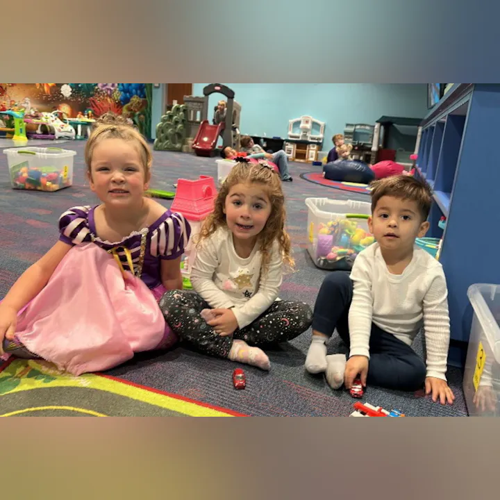 Three children play together on a colorful carpet in a play area, surrounded by toys, with one dressed as a princess.