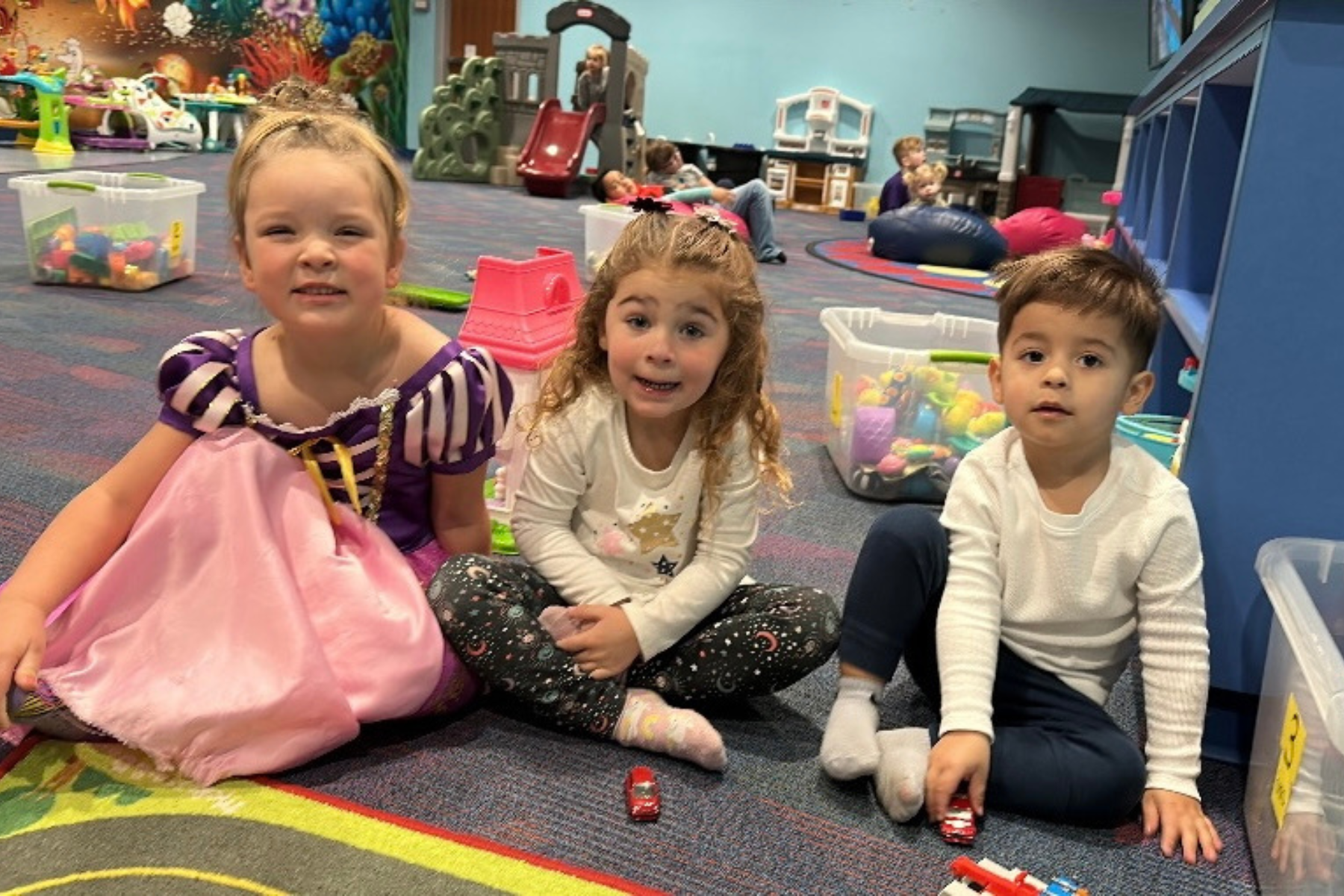 Three children play together on a colorful carpet in a play area, surrounded by toys, with one dressed as a princess.