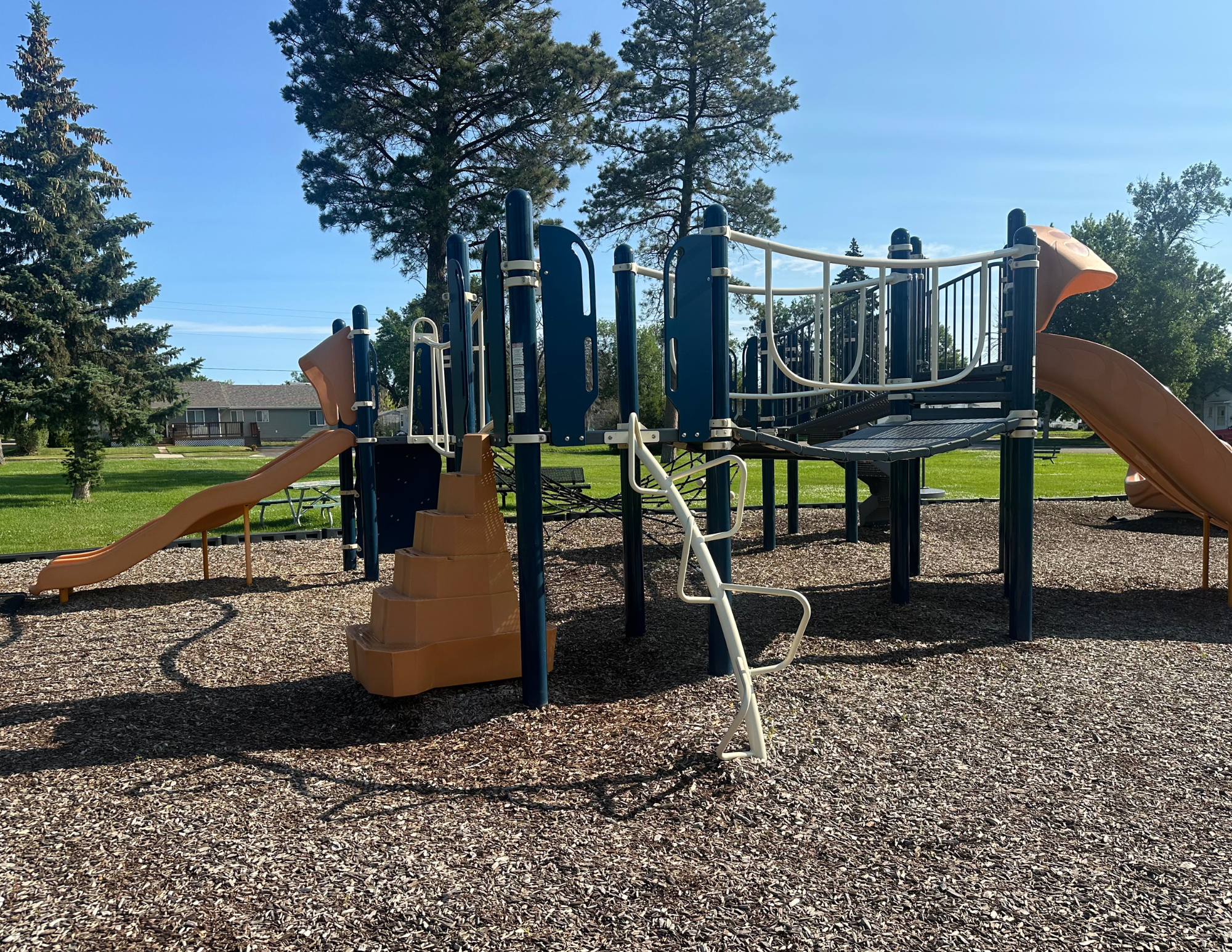 Playground structure with slides, climbing features, and woodchip ground, surrounded by trees and grass.