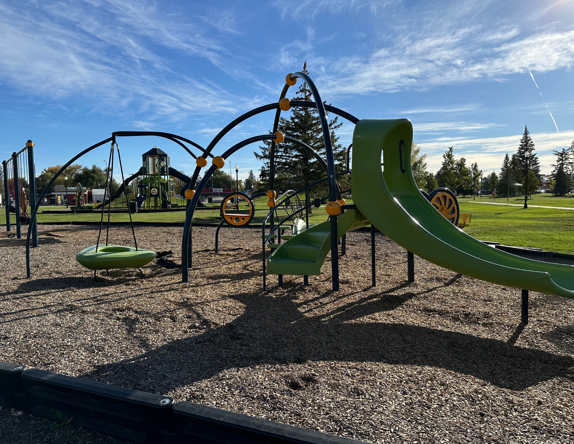 Playground with slides, swings, climbing structures, and wood chip ground cover. Blue sky and trees in the background.