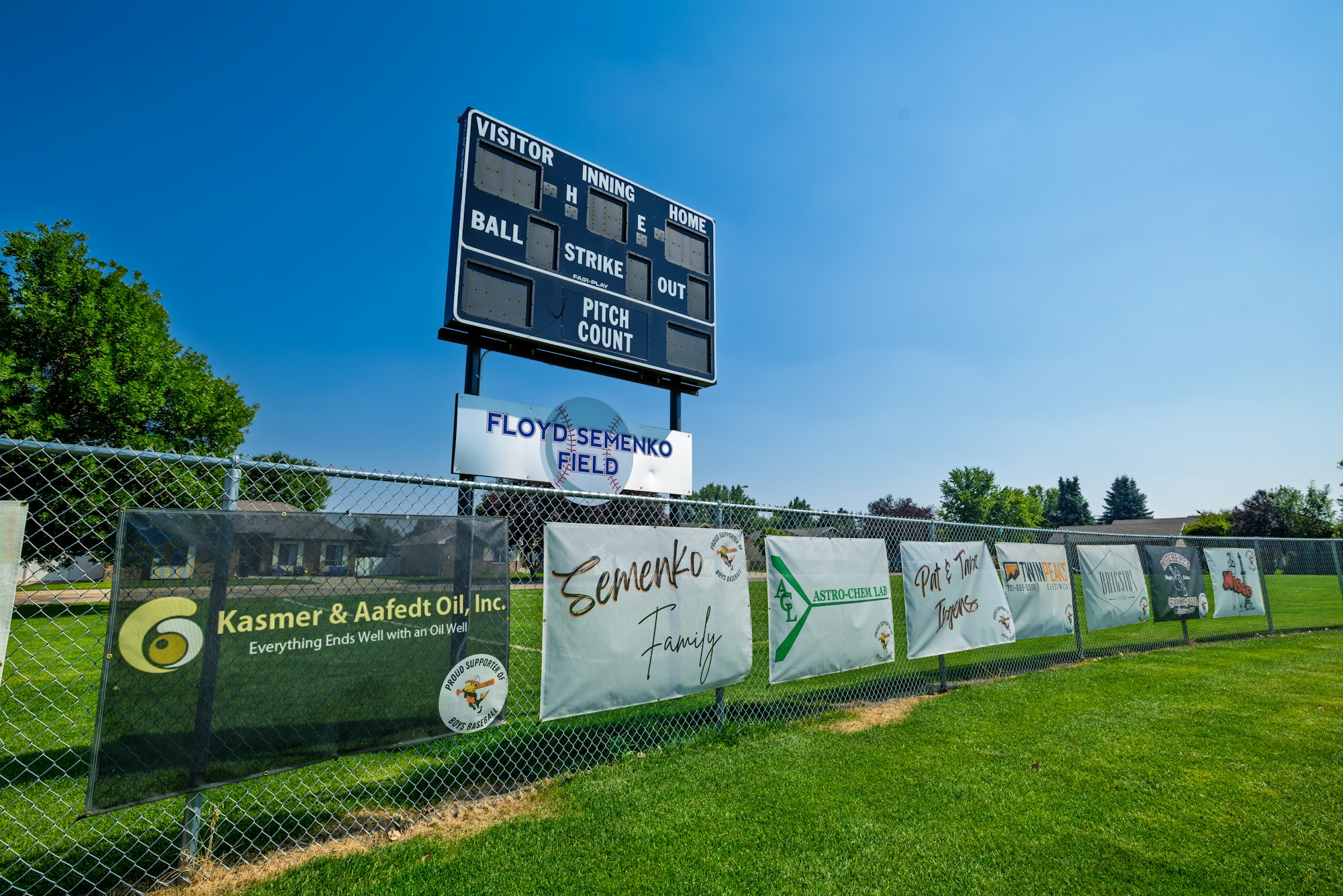 The image shows a baseball field scoreboard and various sponsor banners at Floyd Semenko Field, set against a clear blue sky.