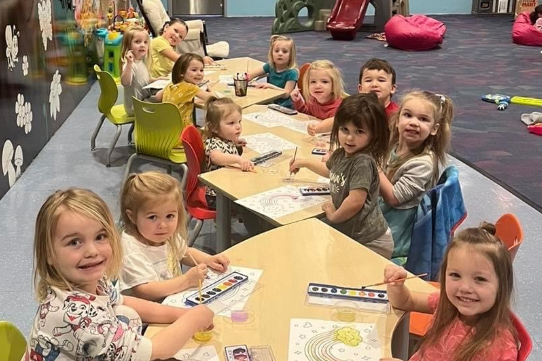Children sitting at tables, engaging in a painting activity in a colorful playroom.