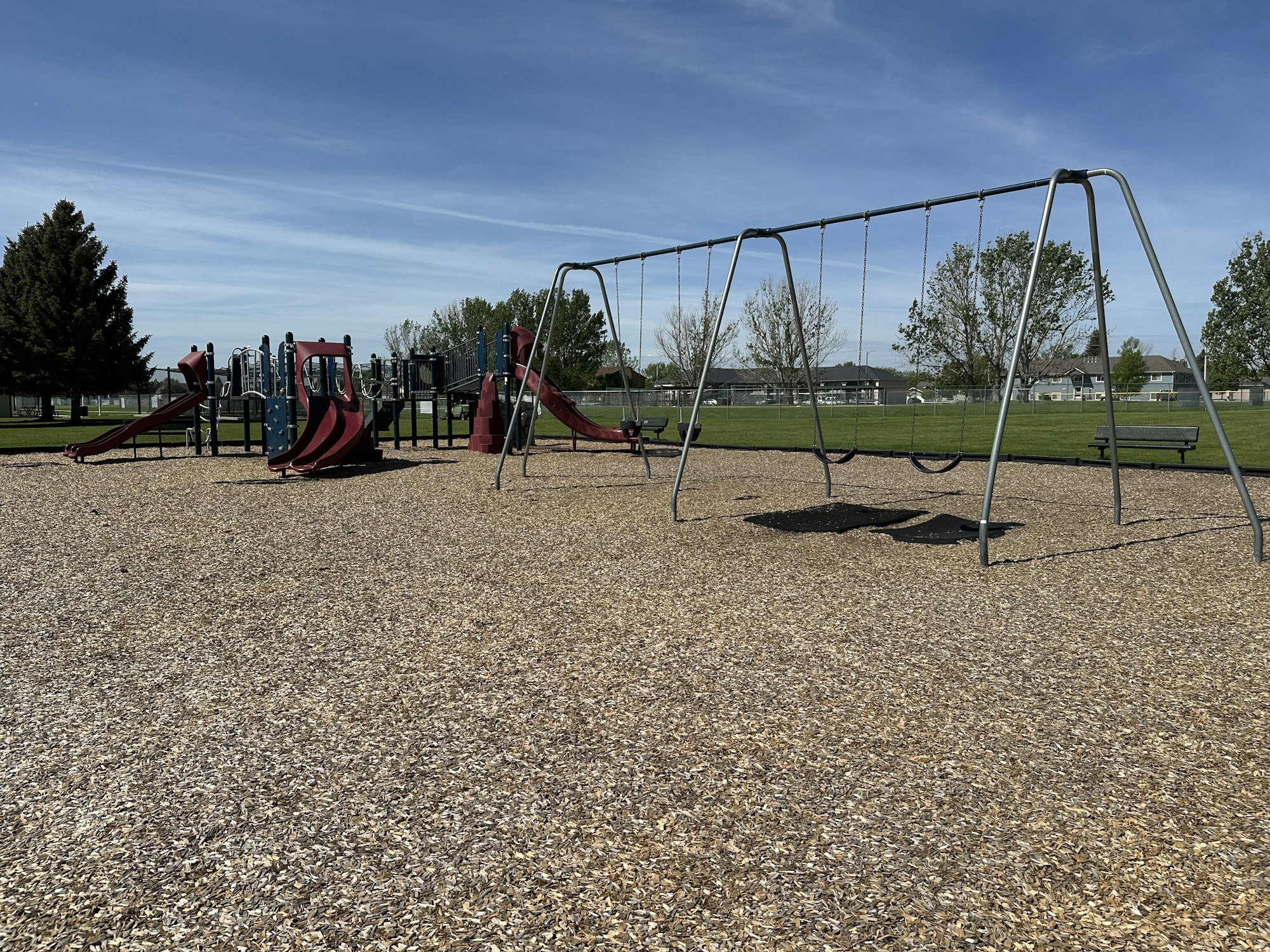Playground with swings, slides, and climbing structures on a sunny day, set on wood chips, with trees and a field in the background.