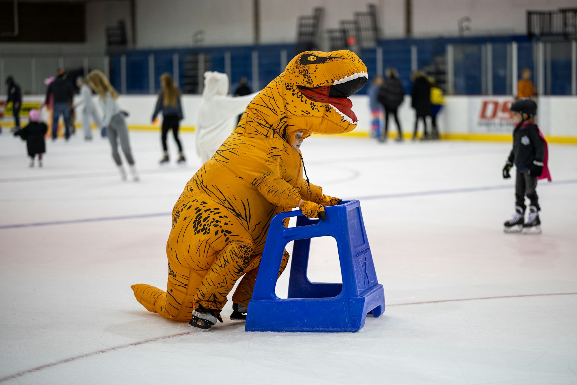A person in a dinosaur costume ice skates while holding onto a blue skating aid, surrounded by others on the rink.