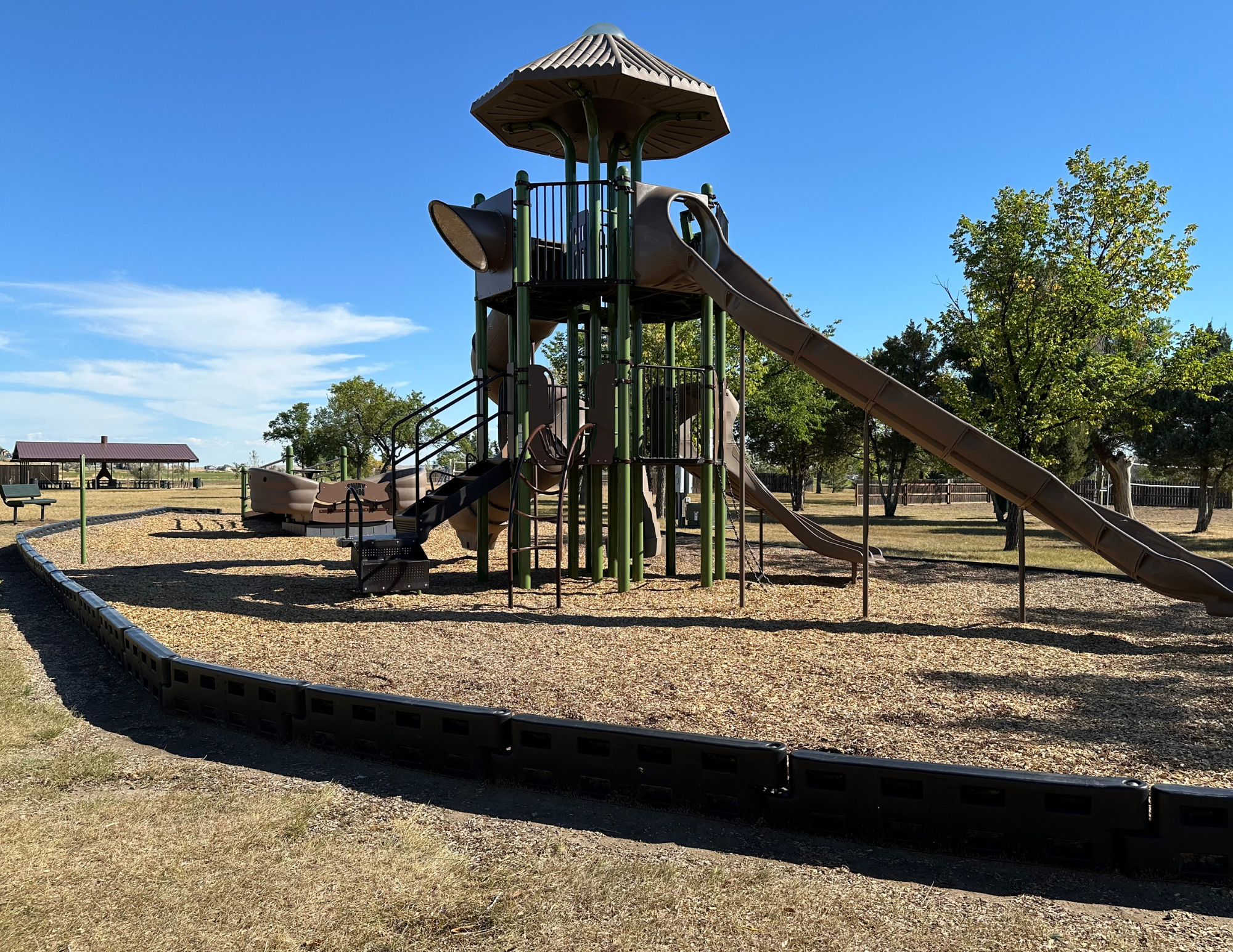 A playground with slides, stairs, and climbing structures on a sunny day. Trees and a pavilion are in the background.