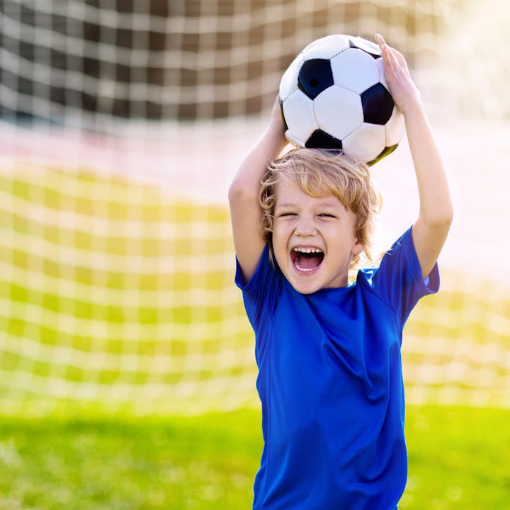 A joyful child in a blue shirt celebrates while holding a soccer ball above his head, with a goal net in the background.