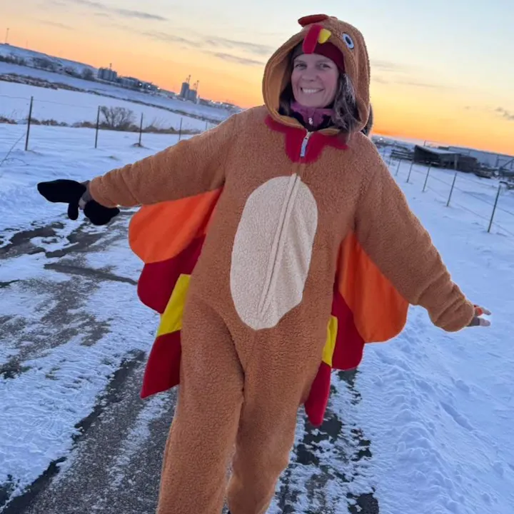 Person in a turkey costume standing on a snowy path with an orange sky at sunset in the background.