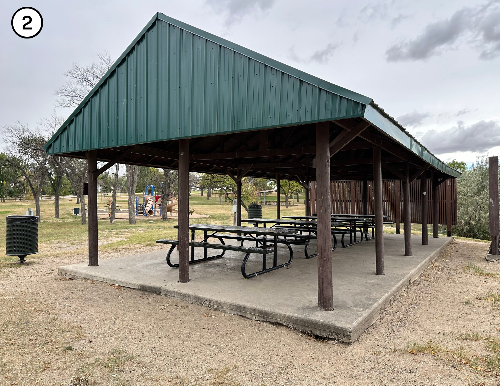 A park pavilion with picnic tables on a concrete slab. There's a playground and trash bins in the background.