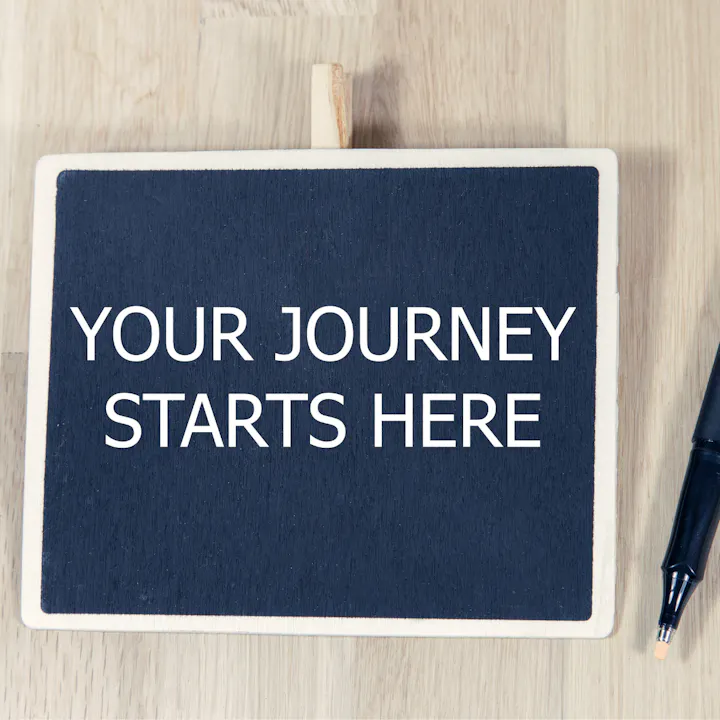 A sign reading "YOUR JOURNEY STARTS HERE" next to a pen on a wooden surface.