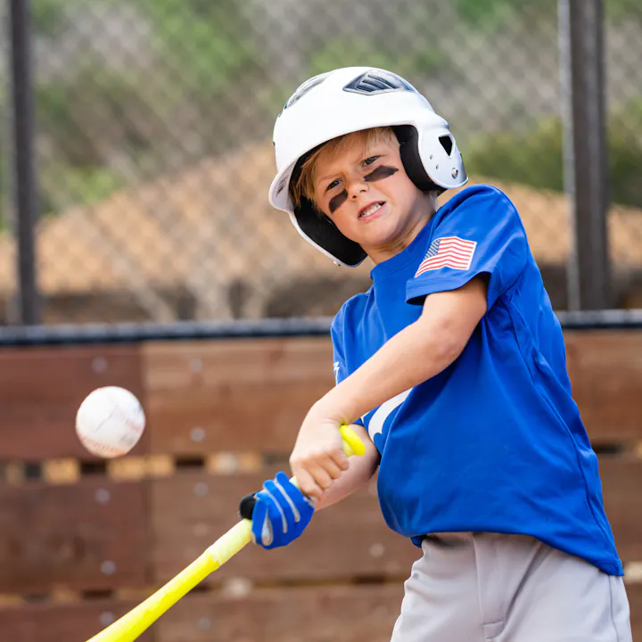 A young boy in a blue shirt and helmet swings a bat at a baseball, showing determination on his face.