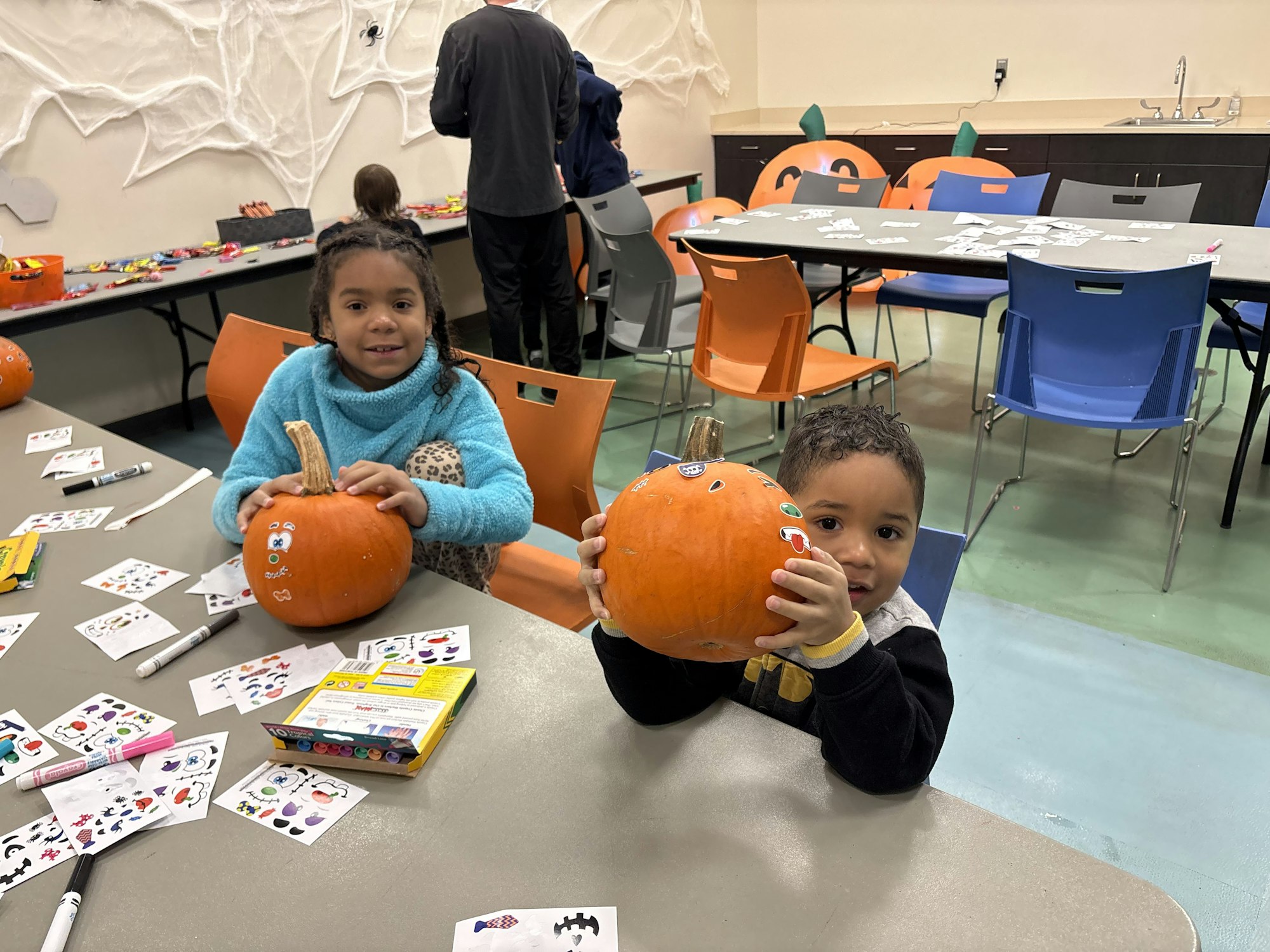 Two children are decorating pumpkins with stickers at a table, surrounded by craft supplies in a festive, decorated space.